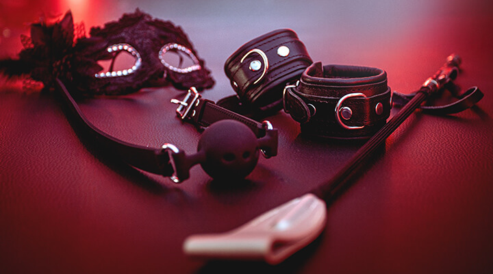 Set aus BDSM‑Accessoires mit Maske, Handfesseln, Ballgag und Reitgerte auf dunkler Oberfläche in rotem Licht.