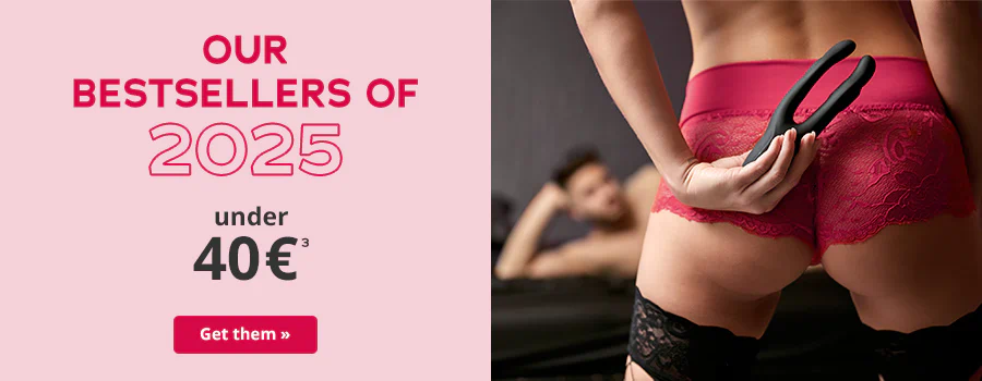 Woman wearing red lace underwear holds a black toy behind her back, with a man lying on a bed in the background. Text: 'Our bestsellers of 2025 – under 40 €'. CTA: 'Get them'