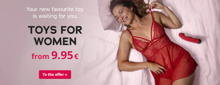A smiling woman is lying on pink bed linen. She is wearing red lace lingerie and a transparent babydoll. A red & black vibrator is next to her hip. Text: “Your new favourite toy is waiting for you”, “TOYS FOR WOMEN from 9.95€” and “To the offer»”.