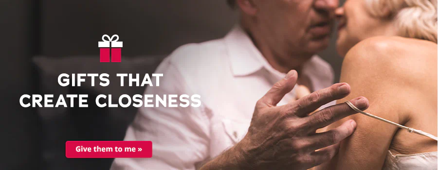 The image shows an advertisement in which a woman and a man are standing close together. The man is wearing a white shirt and the woman is wearing a light-coloured top with thin straps. Their heads are close together and the man is pulling down the thin strap of the woman's top on her shoulder with his hand. The slogan ‘Gifts that create closeness’ is written in large, white letters. A gift is symbolically depicted. Below is a red button with the white inscription ‘Give them to me’.