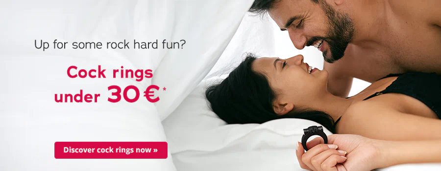 A man and a woman are lying in bed together. They are smiling at each other. The man is topless and the woman is wearing a black top. The woman is holding a black cock ring in her hand. The text: “Up for some rock hard fun? Cock rings under 30 €” is written in large letters. At the bottom is a red button with the text “Discover cock rings now”.