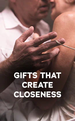 The advertisement has a picture of a woman and a man standing close together. The man is wearing a white shirt and the woman is wearing a light-coloured top with thin straps. Their heads are close together. The man is pulling a thin strap of the woman's top down her shoulder with his hand. The text "Gifts that create closeness" in large, white letters can be seen. There is a red button at the bottom with white text that says "Show them to me". The background is dark.