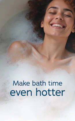 A smiling woman with curly brown hair is lying back, relaxed, in a white bath filled with bubbles. The words "Make bath time even hotter" are displayed prominently. At the bottom, a red button displays the text "Find waterproof toys »".